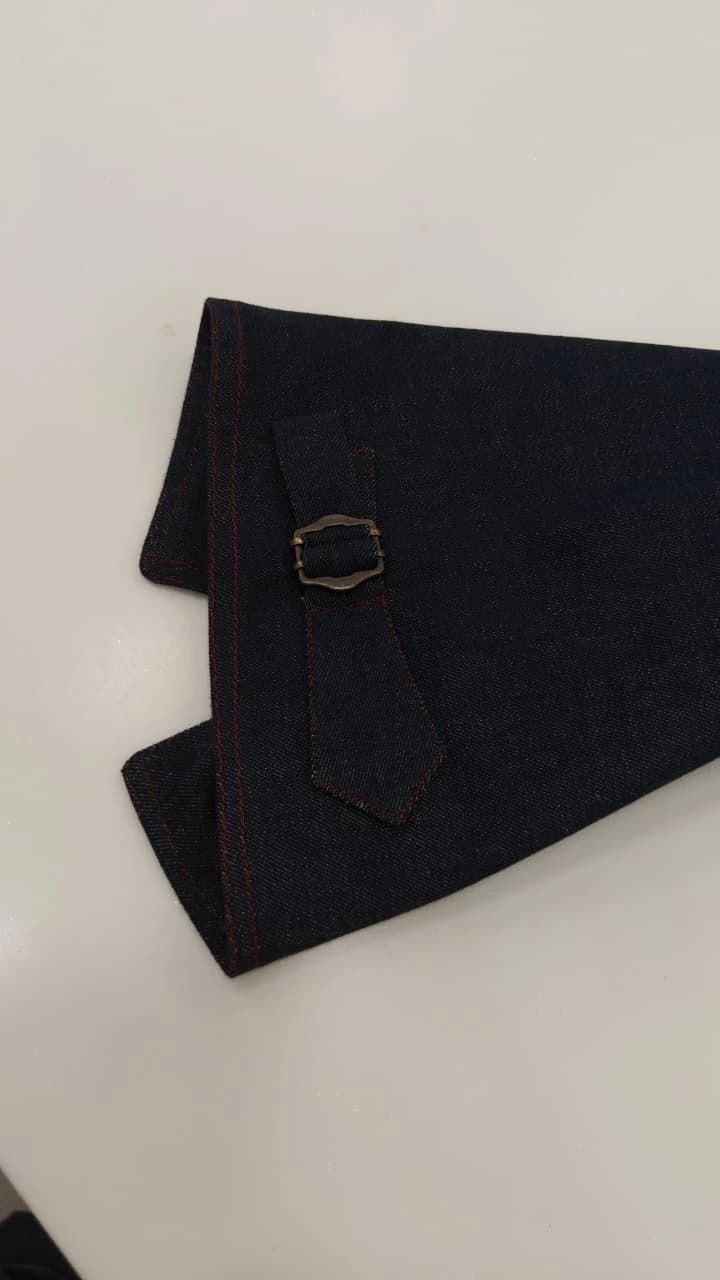A quiet study of a dark denim cuff: red stitching and a small antique brass buckle against an ivory surface.